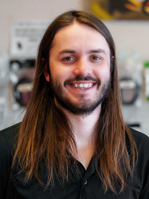 Smiling man with long brown hair and a beard, wearing a black shirt, portrait in an indoor setting with blurred equipment in the background.