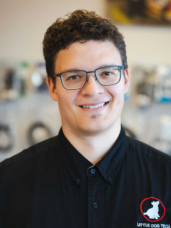Smiling person wearing blue-framed glasses and a black shirt with a red circular patch featuring a white dog silhouette and the text 'Little Dog Tech' on the chest.