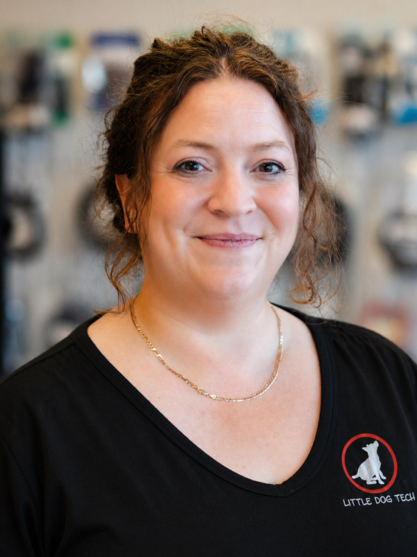Smiling woman in a black Little Dog Tech shirt, standing in a blurred workshop background.