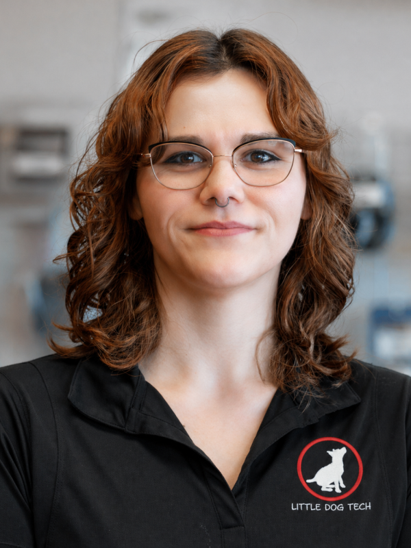 Portrait of a woman with red-brown wavy hair and glasses, wearing a black polo with the Little Dog Tech logo in a workshop setting